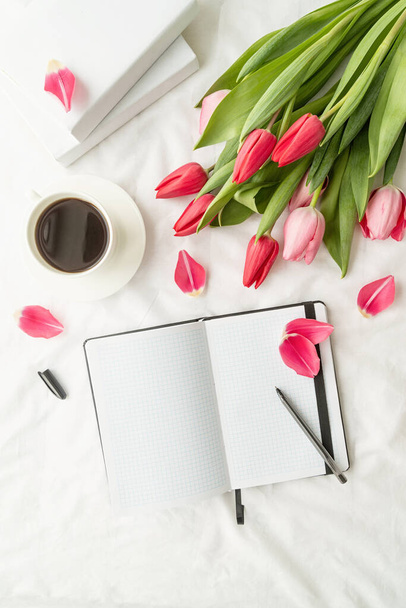 Fiesta y celebración. Vacaciones de primavera. Vista superior del cuaderno en blanco abierto con tulipanes y taza de café en la cama blanca - Foto, Imagen