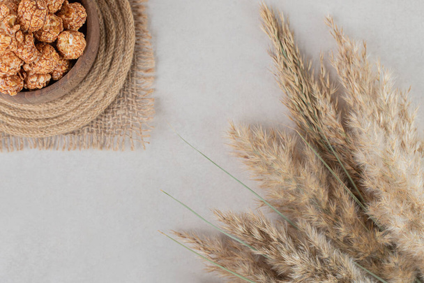 Стебли сушеной травы рядом с деревянной миской на тривету, наполненной леденцовым попкорном на мраморном фоне. Высокое качество фото - Фото, изображение