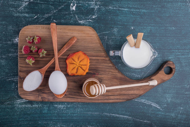 tasse de lait et de baies, tranche de fruits, oeuf et sucre dans des cuillères et miel sur planche de bois - Photo, image
