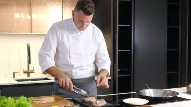 Μαγειρεύοντας ένα νόστιμο πιάτο γαλοπούλα στην κουζίνα του σπιτιού σας. Οι ζουμερές φέτες γαλοπούλας με σκόρδο και δεντρολίβανο τηγανίζονται σε γκριλ. Ένας άντρας με ποδιά γυρίζει το ψητό κρέας με λαβίδες. Γαλακτοκομικά - Πλάνα, βίντεο