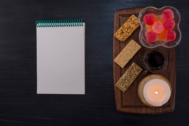 blank notebook, snack bars, candle, tea and sweets in glass on wooden board - Photo, image