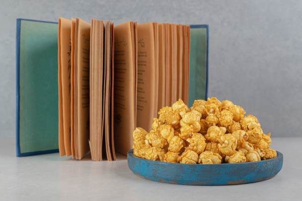 Open book next to an overfilled small tray of caramel popcorn on marble background. High quality photo - Photo, Image