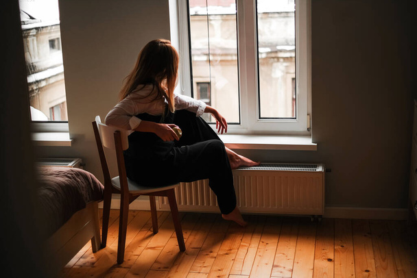 όμορφη κοπέλα με μαύρο παντελόνι κάθεται σε μια καρέκλα και κοιτάζει έξω από το παράθυρο στο δωμάτιό της. μοντέρνο εσωτερικό διαμέρισμα - Φωτογραφία, εικόνα