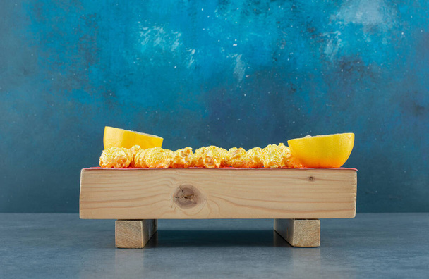 Wooden board filled with crunchy caramel coated popcorn bits and sliced lemon pieces on blue background. High quality photo - Photo, Image