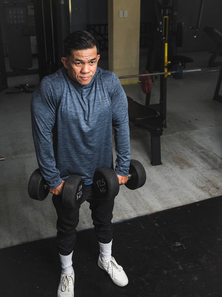 A buff asian guy does dumbbell shrugs at the gym. Traps workout and exercise. - Photo, Image