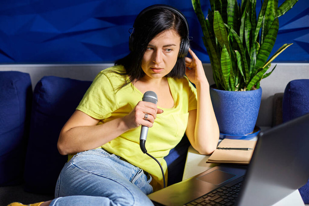 Χαρούμενη γυναίκα σε casual ρούχα Ηχογράφηση ένα Podcast, μιλώντας σε ένα μικρόφωνο με ακουστικά και laptop, σημειωματάριο, Γυναίκα podcaster ζωντανή ροή, ή τραγουδώντας τραγούδι karaike. - Φωτογραφία, εικόνα