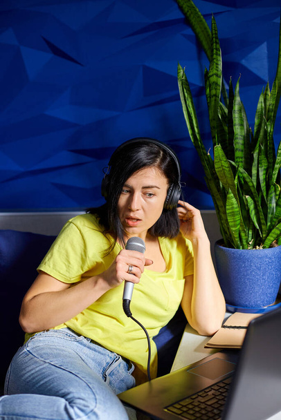 Vrolijke vrouw in casual kleding Opname van een podcast, praten in een microfoon met koptelefoon en laptop, notebook, vrouwelijke podcaster live streaming, of zingen karaike lied. - Foto, afbeelding