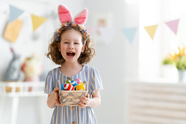 Schönes Kind mit bemalten Eiern. Glückliche Familie bereitet sich auf Ostern vor. Nettes kleines Mädchen trägt Hasenohren.   - Foto, Bild