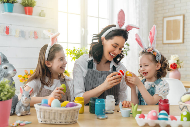 Happy holiday! A mother and her daughters are painting eggs. Family preparing for Easter. Cute little children girls are wearing bunny ears. - Fotografie, Obrázek
