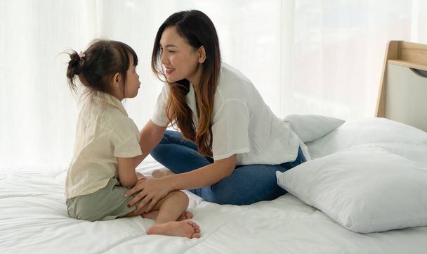 азиатская мама и молодой ребенок с улыбающимся лицом, сидящие вместе на кровати в спальне - Фото, изображение