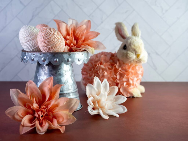 Easter Bunny Decor sitting by a Raised Rustic Metal Dish Filled with String Decorated Easter Eggs surrounded by peach artificial dahlia flower  blooms, perfect for the spring holiday! - Photo, Image