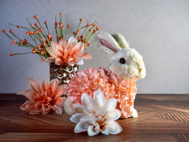 Decoración de conejo de Pascua sentado en una superficie de madera rodeada de flores de dalia artificial melocotón, perfecto para las vacaciones de primavera! - Foto, Imagen