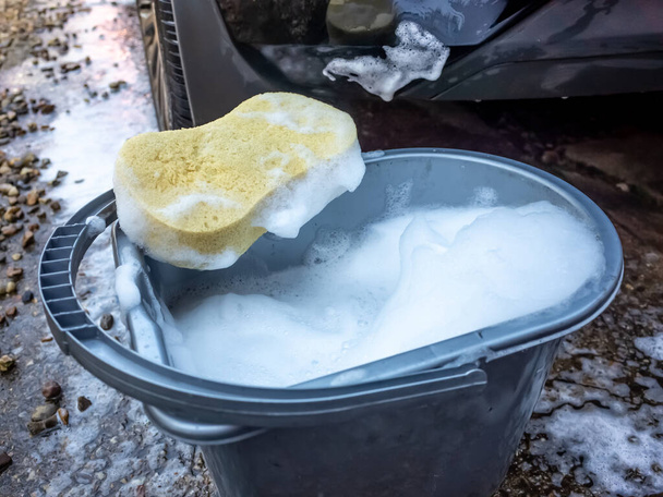 Gele spons op een emmer zeepwater voor een vuile achterbumper van een auto - Foto, afbeelding