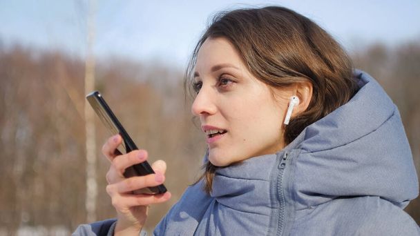 Młoda kobieta ze słuchawką bezprzewodową używa smartfona, by wysłać wiadomość głosową do nowego klubu społecznościowego, stojącego gdzieś na zewnątrz parku. - Zdjęcie, obraz