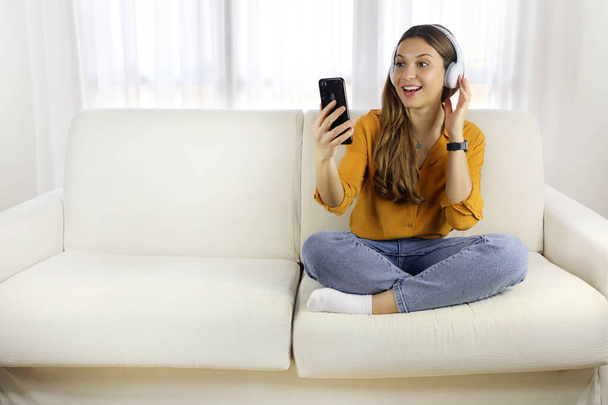 Ενθουσιασμένη νεαρή γυναίκα κάθεται με τα πόδια σταυρωμένα στον καναπέ βλέποντας μουσική βίντεο στο smartphone στο σπίτι. Αντιγραφή χώρου. - Φωτογραφία, εικόνα