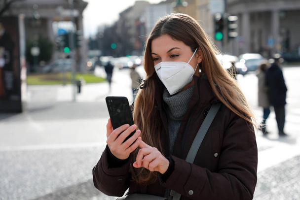 Πορτρέτο της κυρίας σε χειμωνιάτικα ρούχα και FFP2 KN95 μάσκα προσώπου με τα πόδια στο δρόμο της πόλης πληκτρολογώντας στο smartphone - Φωτογραφία, εικόνα