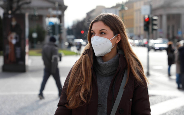 Αίσθηση σύγχυσης. Νεαρή γυναίκα με χειμωνιάτικα ρούχα που περπατάει στο δρόμο φορώντας προστατευτική μάσκα FFP2 KN95. Κορίτσι με μάσκα προσώπου αισθάνεται μόνο κατά τη διάρκεια μιας πανδημίας. - Φωτογραφία, εικόνα