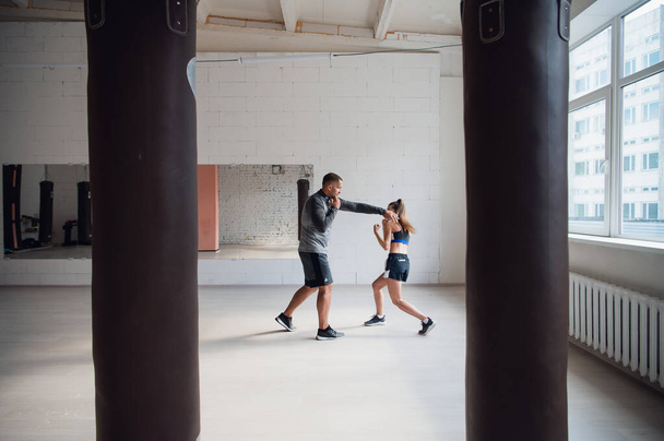 Plan moyen de la jeune femme faisant de l'entraînement de boxe au gymnase et frappant son instructeur, qui esquive ses coups et contrôle le processus d'entraînement. - Photo, image