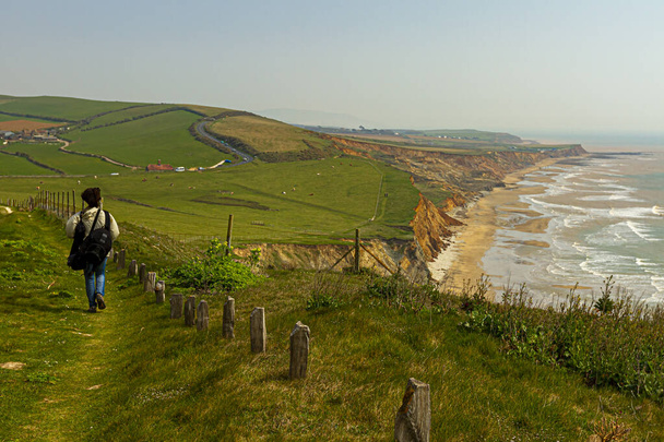 Idyllic coast of Isle of Wight with cliffs, Atlantic Ocean, shoreline, pastures and farms at a distance and a woman hiker with backpack walking alone on a trail path. A rural scenery, natural beauty. - Photo, Image