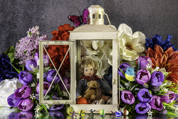 кукла и плюшевый мишка в металлическом фонаре, отражающемся на стеклянном зеркале в окружении весенних цветов - Фото, изображение