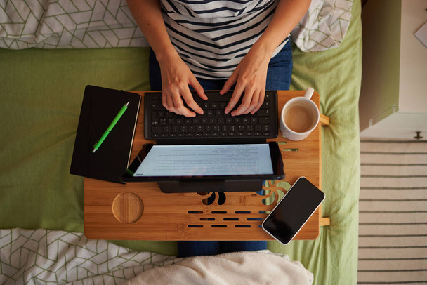 Top view of a woman telecommuting from her bed. He types on her laptop, with a mug and notebook on the table. The room has a decorative wooden wall. - Photo, Image