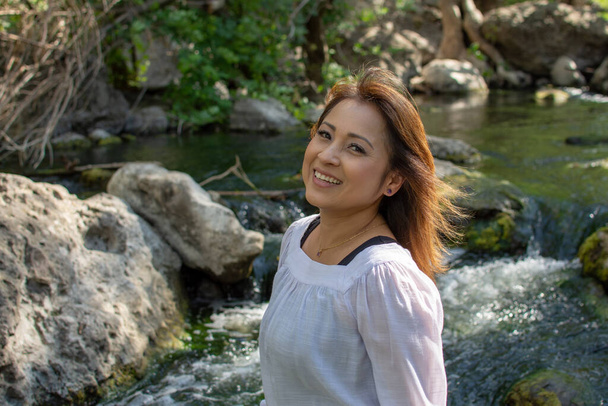 Латиноамериканка с улыбкой, стоящая в тени с светящимися волосами в ручье с водопадами на заднем плане весной - Фото, изображение
