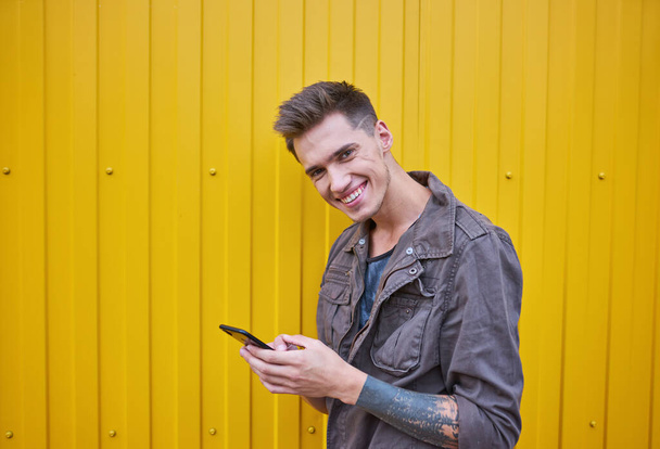 young guy stands near the yellow wall, looks into the phone and smiles. - Photo, Image