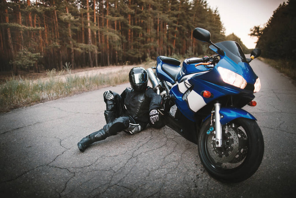 Motociclista con un traje de protección y un casco negro se sienta junto a su motocicleta en la carretera. Motociclista descansando cerca de una motocicleta - Foto, Imagen