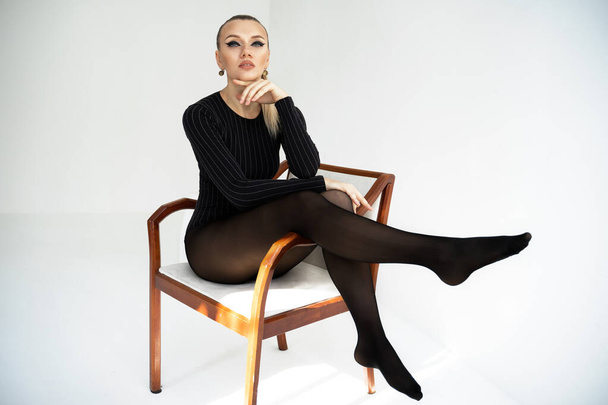 Modell in schwarzen Strumpfhosen auf weißem Hintergrund. heiße blonde Mädchen mit langen Beinen in Dessous sitzen auf dem Stuhl und beobachten uns.  - Foto, Bild