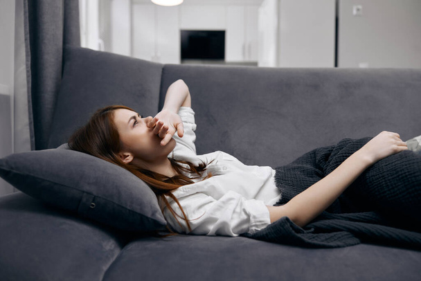 Женщина, держащая руку на лице, лежала на диване, прячась за одеялом, чувствуя себя нездоровой. - Фото, изображение