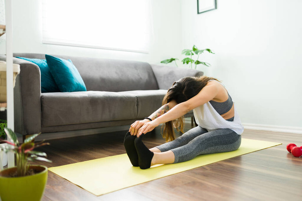Giovane donna ispanica che fa una piega seduta in avanti durante una routine di yoga. Donna attiva che lavora con un tappetino da ginnastica a casa - Foto, immagini