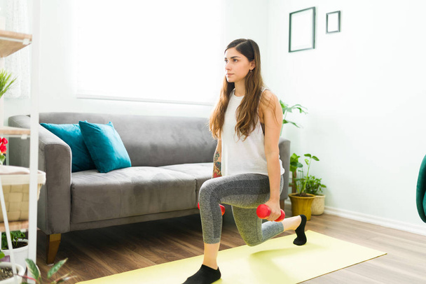 Deportiva latina haciendo saltos de piernas en la sala de estar mientras sostiene dos pesas. Mujer con un estilo de vida saludable haciendo ejercicio en casa - Foto, imagen