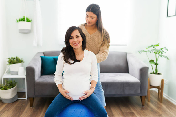 Portrait of a beautiful happy pregnant woman smiling while sitting on a fitness ball and receiving a back massage from her hispanic doula - Photo, Image