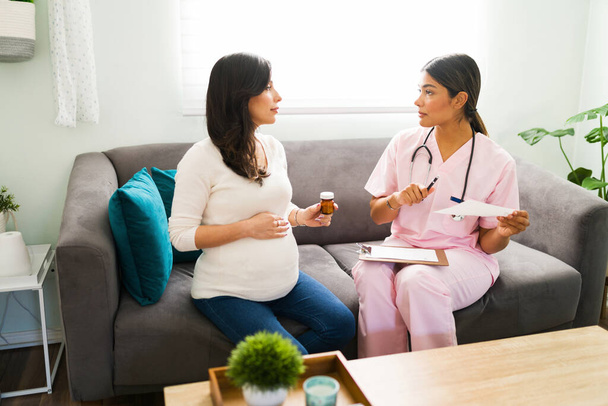 Vonzó női orvos orvosi receptet és egy üveg tablettát ad egy csinos terhes nőnek, miközben a kanapén ül. - Fotó, kép