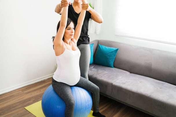 Mulher muito grávida trabalhando fora e fazendo exercícios de alongamento com uma bola de fitness na sala de estar com a ajuda de sua doula  - Foto, Imagem