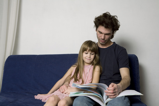 Father and daughter are sitting on the sofa and reading a book - Photo, image