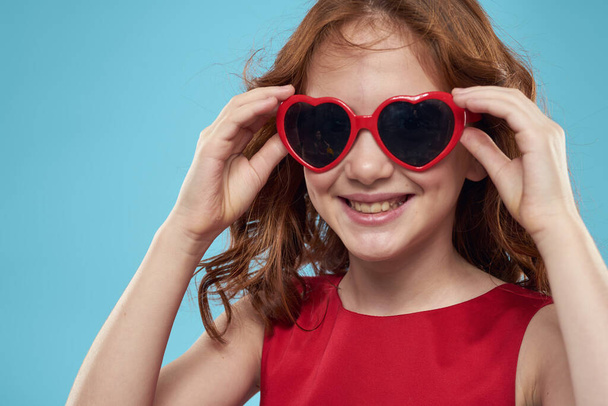 Little girl in sunglasses in the form of hearts red dress curly hair blue background - Photo, Image