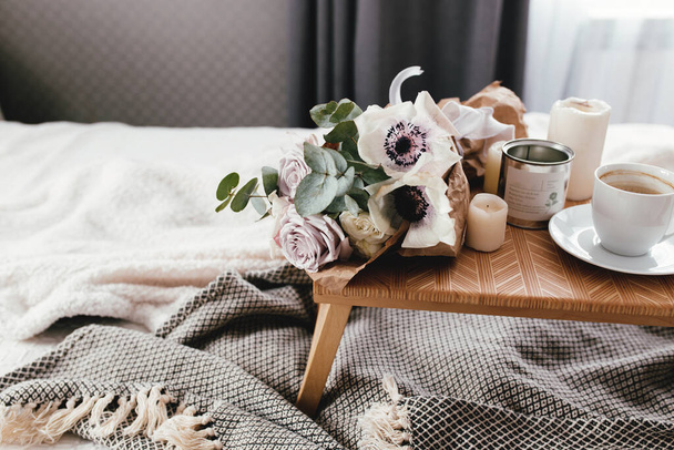 Ρομαντικό πρωινό. Ξύλινο τραπεζάκι σαλονιού με λουλούδια στο κρεβάτι με καρό, φλιτζάνι καφέ, λουλούδια και κεριά. Λιλά τριαντάφυλλα με ευκάλυπτο και ανεμώνες. Εσωτερικοί γκρι τόνοι. Υψηλής ποιότητας φωτογραφία - Φωτογραφία, εικόνα
