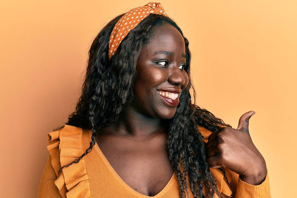 Afrikaanse jonge vrouw dragen casual kleding over gele achtergrond glimlachen met gelukkig gezicht kijken en wijzen naar de zijkant met duim omhoog.  - Foto, afbeelding
