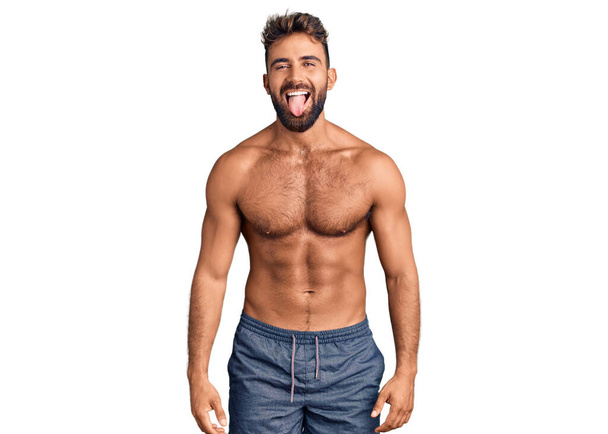 Junger hispanischer Mann in Badebekleidung, der die Zunge mit lustigem Gesichtsausdruck herausstreckt. Emotionales Konzept.  - Foto, Bild