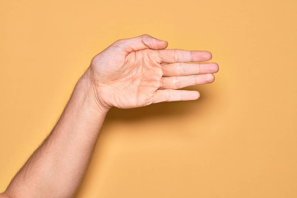Χέρι καυκάσιου νεαρού άνδρα που δείχνει τα δάχτυλά του πάνω από απομονωμένο κίτρινο φόντο που εκτείνεται και φτάνει με ανοιχτό χέρι για χειραψία, δείχνοντας παλάμη - Φωτογραφία, εικόνα