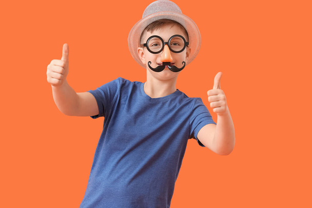 Little boy in funny disguise showing thumb-up gesture on color background. April Fools Day celebration - Foto, Imagem