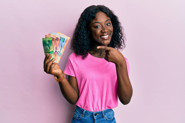 вродлива африканська молода жінка, що тримає південноафриканські банкноти, усміхається, показуючи пальцем і рукою.  - Фото, зображення