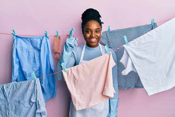 Mujer afroamericana con cabello trenzado lavando ropa en tendedero sonriendo asombrada y sorprendida y señalando con los dedos y los brazos levantados.  - Foto, Imagen