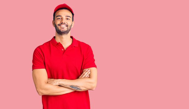 Young handsome hispanic man wearing delivery courier uniform happy face smiling with crossed arms looking at the camera. positive person.  - Photo, Image