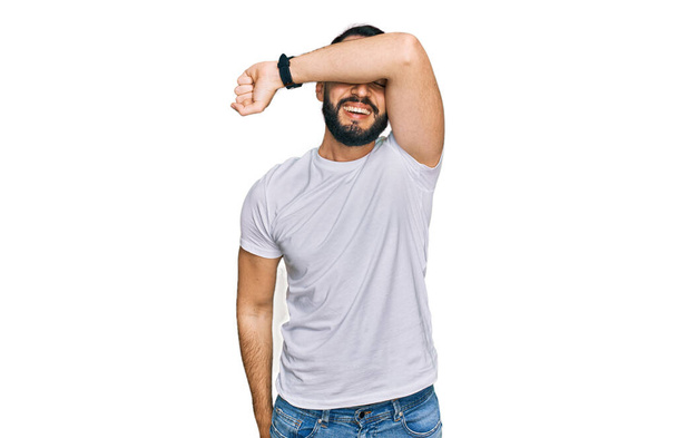 Young man with beard wearing casual white t shirt covering eyes with arm smiling cheerful and funny. blind concept.  - Photo, Image