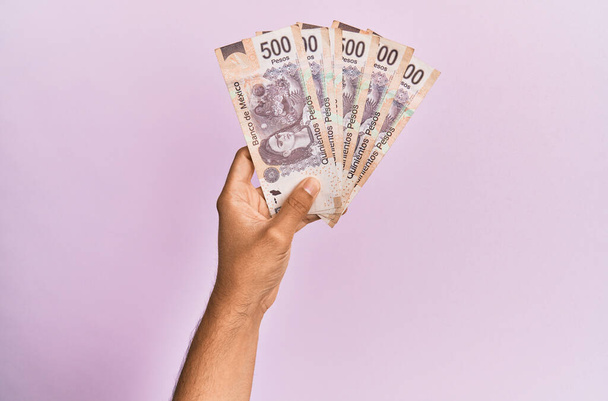 Іспанська рука, що тримає 500 мексіканських песо банкнот над ізольованим рожевим фоном. - Фото, зображення
