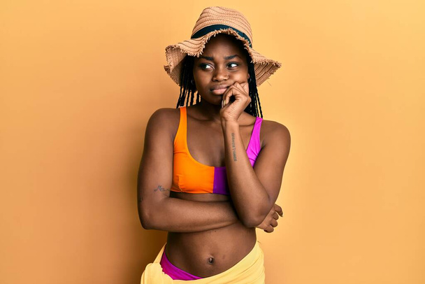 Mladá africká americká žena v bikinách a letním klobouku myšlení soustředěné o pochybách s prstem na bradě a vzhlíží zvědavý  - Fotografie, Obrázek