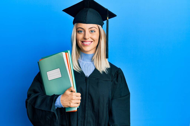 Красива блондинка в шапці для випускних і церемонії одягу тримає книги, які виглядають позитивними і щасливими, стоячи і посміхаючись з впевненою посмішкою, показуючи зуби
  - Фото, зображення