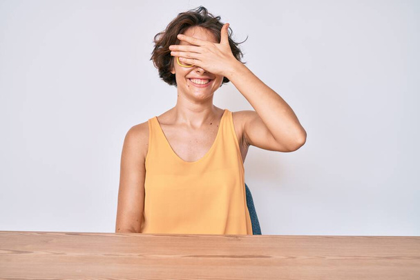 Jonge Spaanse vrouw in casual kleding en een bril zittend op de tafel glimlachend en lachend met de hand op het gezicht ogen bedekkend voor verrassing. blind concept.  - Foto, afbeelding